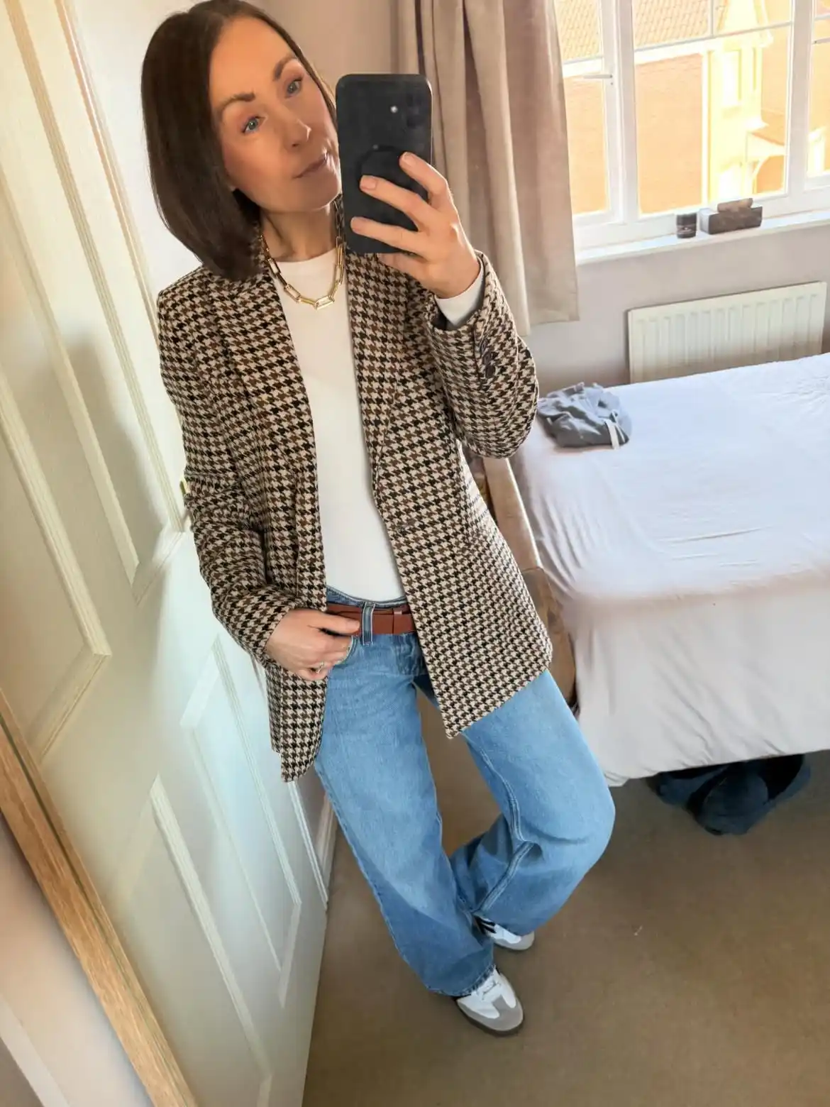 6. Houndstooth Blazer with White Tee and Light Wash Jeans

A brown and cream houndstooth blazer adds instant pattern to this casual outfit. Beneath it, a plain white top keeps the focus on the printed layer above. A chunky gold chain necklace rests near the collar, adding a refined touch. Light wash straight jeans and a slim brown belt bring balance to the top half.

White sneakers finish the look with a relaxed, everyday feel. Pattern mixing feels less daunting when paired with denim and simple basics like this. Weekend brunches or casual office days both suit this outfit, since it blends comfort with just enough structure to feel intentional.